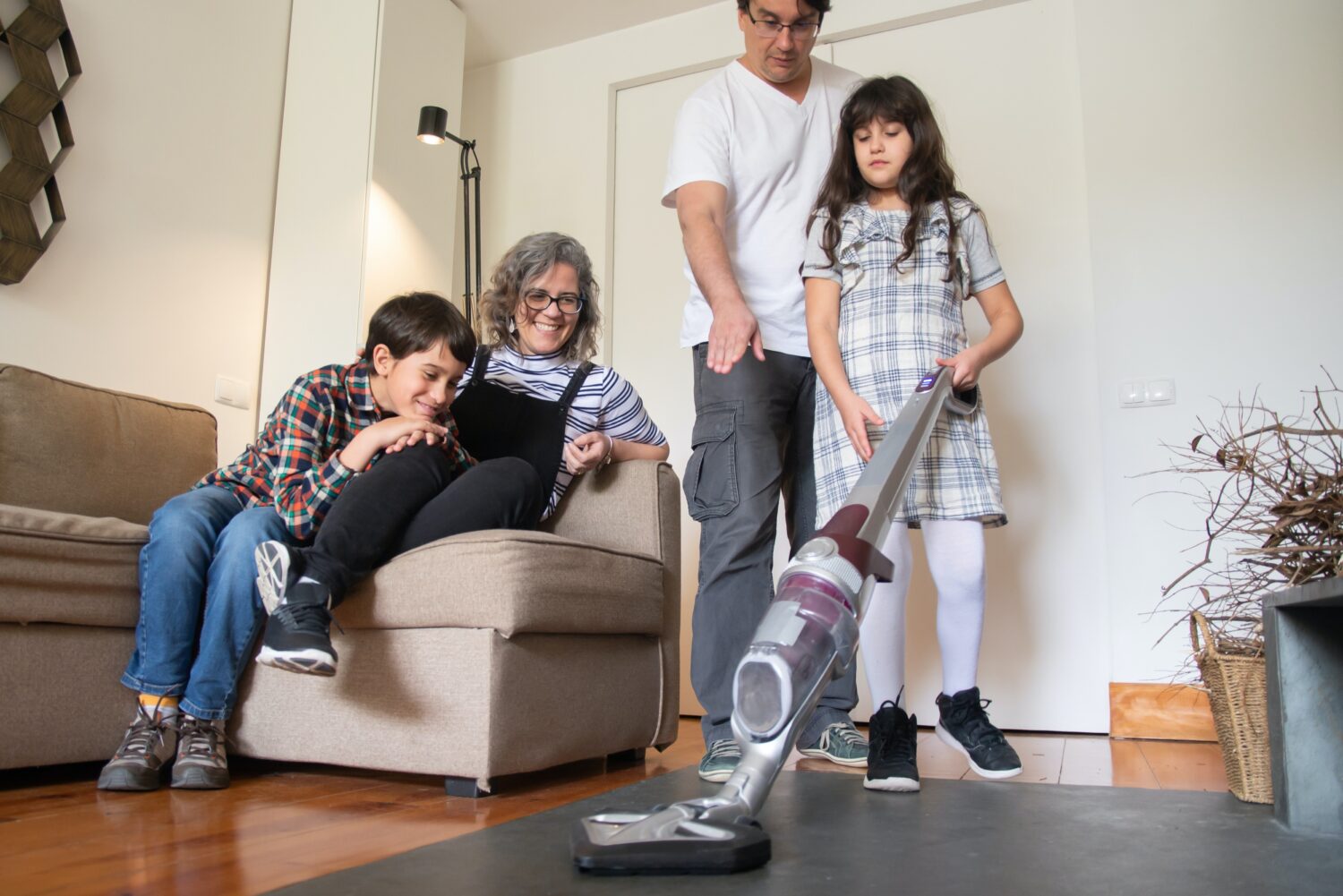 A young girl practices using a bagless vacuum cleaner while her family looks on.
